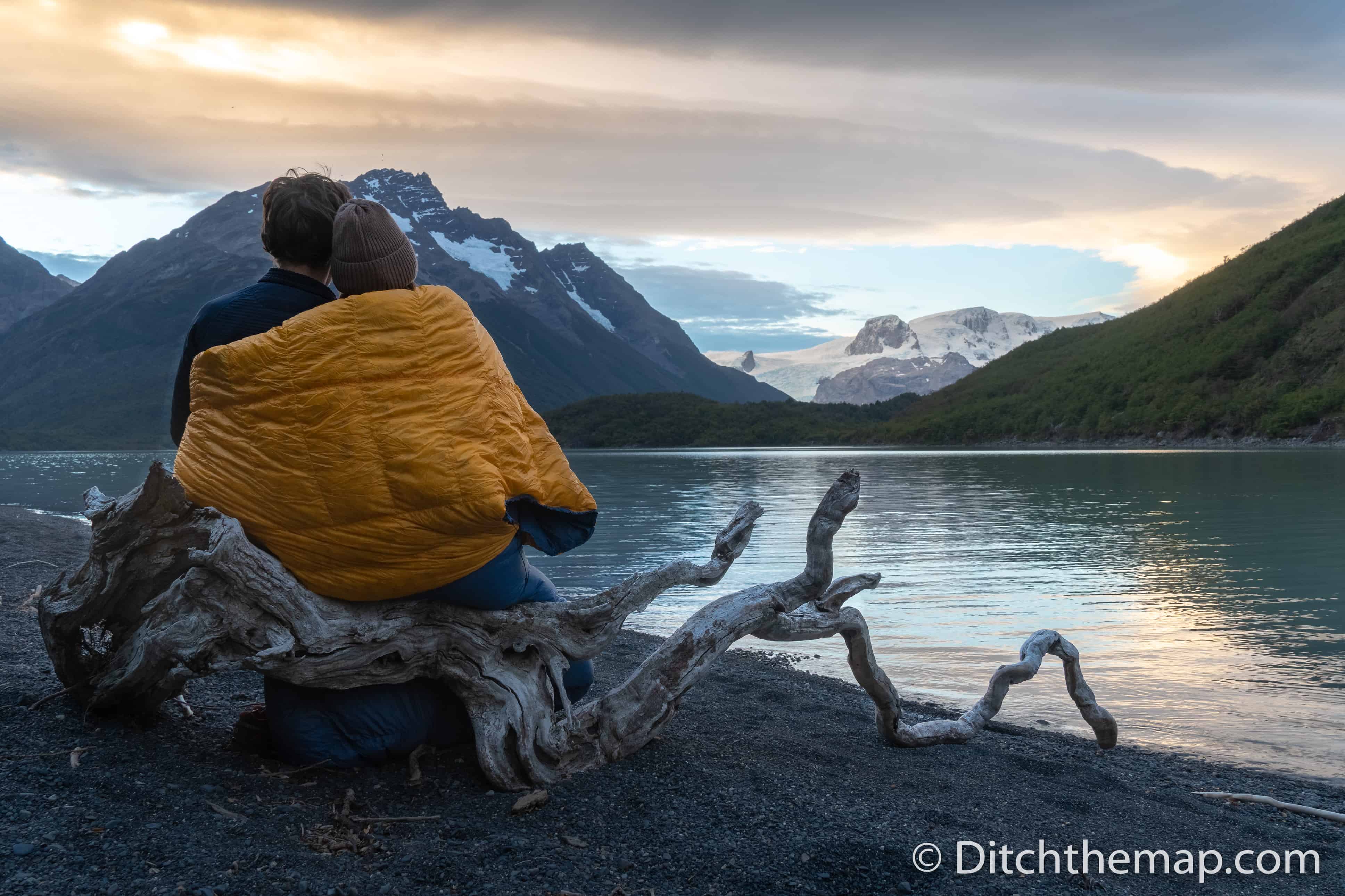 A couple keeping warm with a sleeping bag next to a cold lake and mountain view