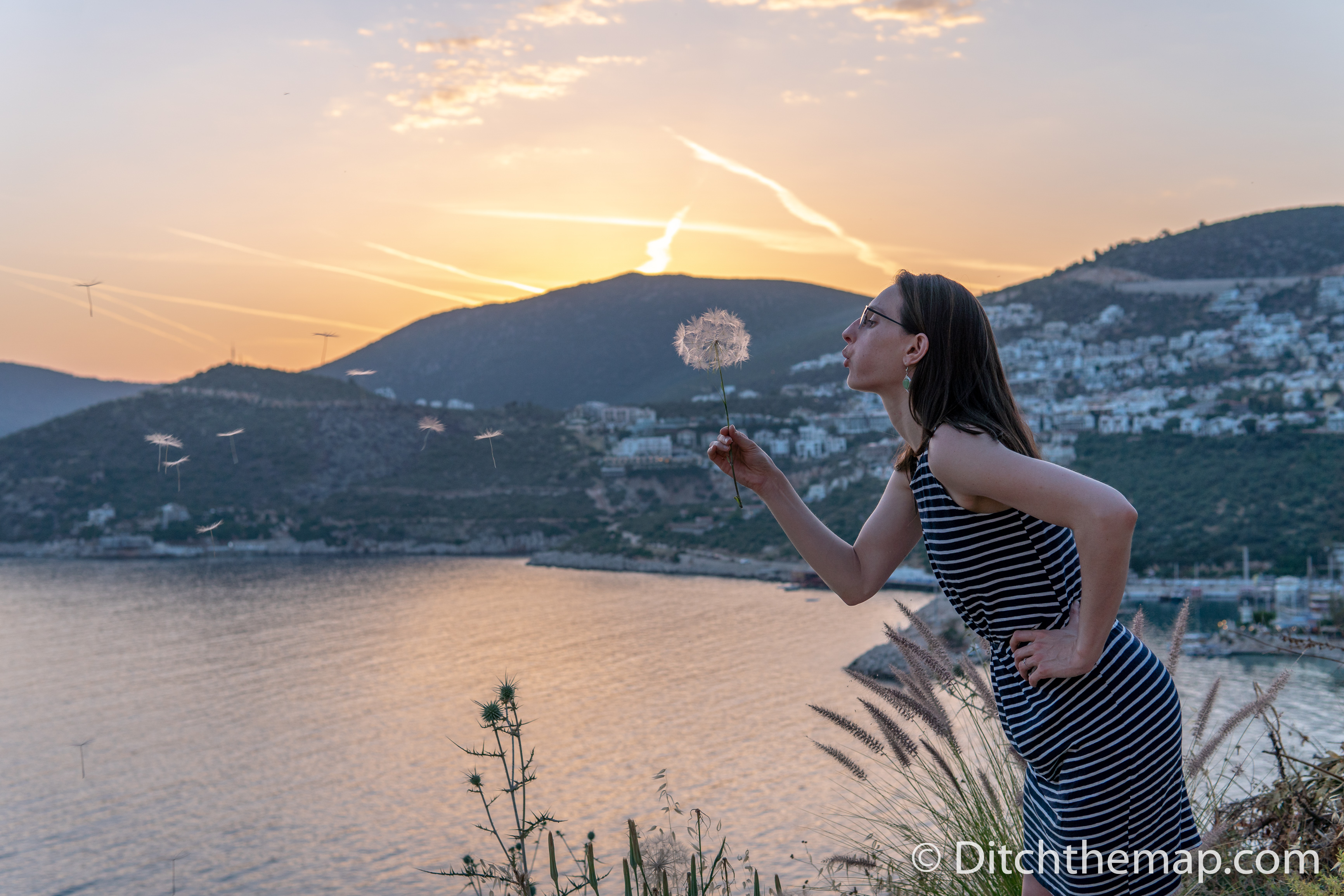 A girl blowing a dandelion during sunset next to a lake and mountains