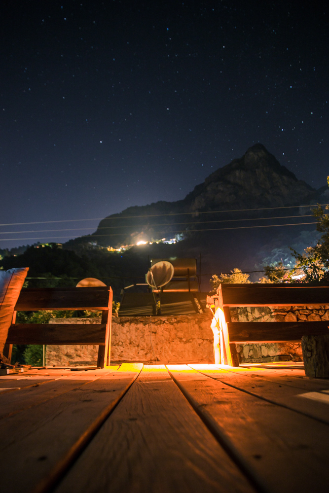 View from the wooden rooftop area at night. Clear sky with lots of stars, cozy warm light from the rooftop, view towards the mountain.